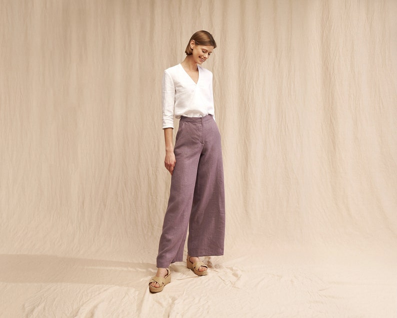 May include: A woman wearing a white button-down shirt and wide-leg pants in a light purple color. She is standing on a light beige background.