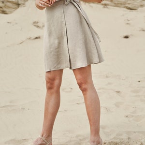 May include: A woman wearing a white tank top and a beige linen wrap skirt. She is standing on a sandy beach and wearing beige platform sandals.