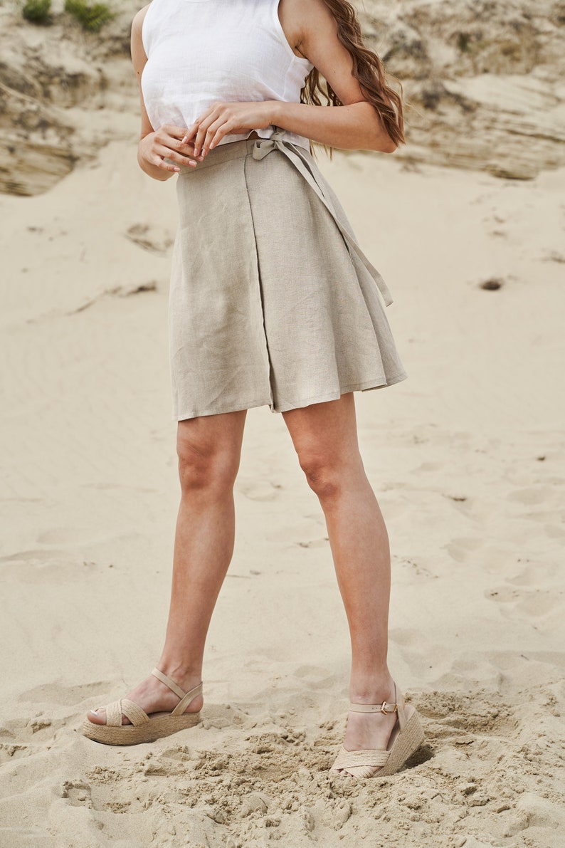 May include: A woman wearing a white tank top and a beige linen wrap skirt. She is standing on a sandy beach and wearing beige platform sandals.