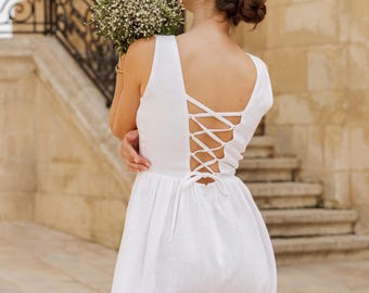 May include: A white linen dress with a lace-up back detail. The dress has a simple, elegant design and is perfect for a summer wedding or other special occasion.