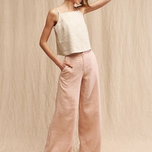 May include: A woman wearing a light beige linen tank top and wide-leg pants in a pale pink color. She is standing on a beige background and smiling.