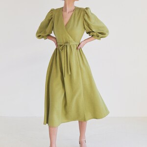 Puede incluir: Un vestido cruzado verde claro con cuello en V y mangas abullonadas hasta el codo. El vestido tiene una cintura anudada y una falda midi. La modelo lleva sandalias rosa claro. El vestido es de lino.