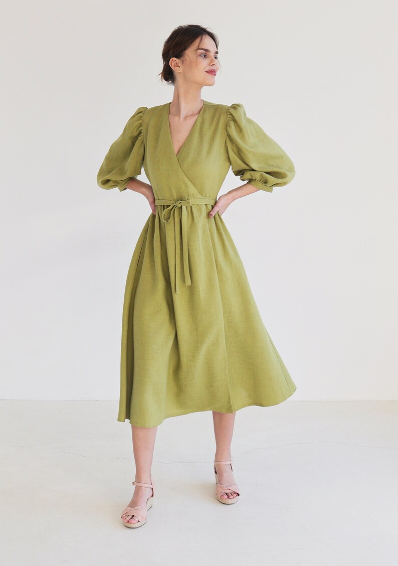 Puede incluir: Un vestido cruzado verde claro con cuello en V y mangas abullonadas hasta el codo. El vestido tiene una cintura anudada y una falda midi. La modelo lleva sandalias rosa claro. El vestido es de lino.