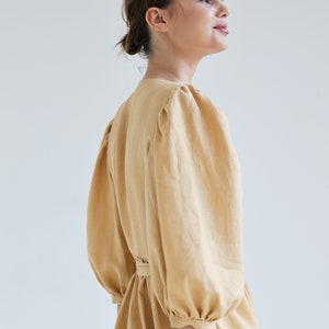 Puede incluir: Vestido amarillo claro con mangas voluminosas y cintura ce&ntilde;ida. El vestido tiene un escote en V y una textura sutil, que sugiere lino o tejido similar. Las mangas est&aacute;n recogidas en los hombros y pu&ntilde;os, creando un efecto abullonado.