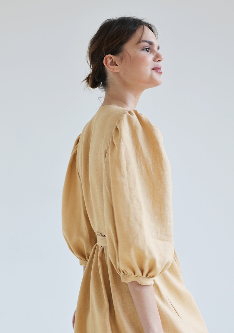 Puede incluir: Vestido amarillo claro con mangas voluminosas y cintura ce&ntilde;ida. El vestido tiene un escote en V y una textura sutil, que sugiere lino o tejido similar. Las mangas est&aacute;n recogidas en los hombros y pu&ntilde;os, creando un efecto abullonado.