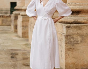 May include: A white linen wrap dress with a V-neckline and puff sleeves. The dress is midi length and has a tie belt at the waist. The model is wearing the dress with white heels.