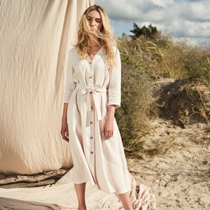 May include: A woman wearing a light beige linen dress with a belt and buttons. She is standing on a sandy beach with a beige fabric backdrop.