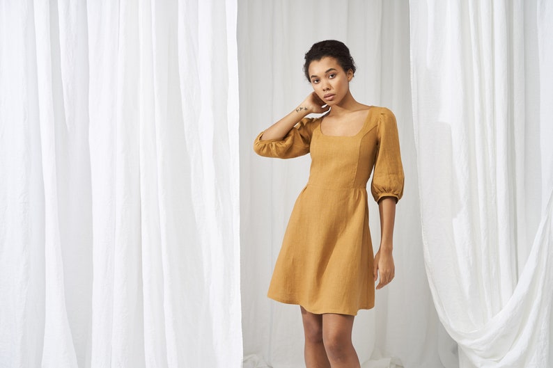 May include: A woman wearing a mustard yellow linen dress with a square neckline and puffed sleeves. The dress has a fitted bodice and a flowy skirt. She is standing in front of a white curtain.