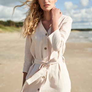 May include: A woman wearing a light beige linen dress with a button-up front and a tie belt. The dress has a v-neck and 3/4 sleeves. The woman is standing on a sandy beach with her hair blowing in the wind.