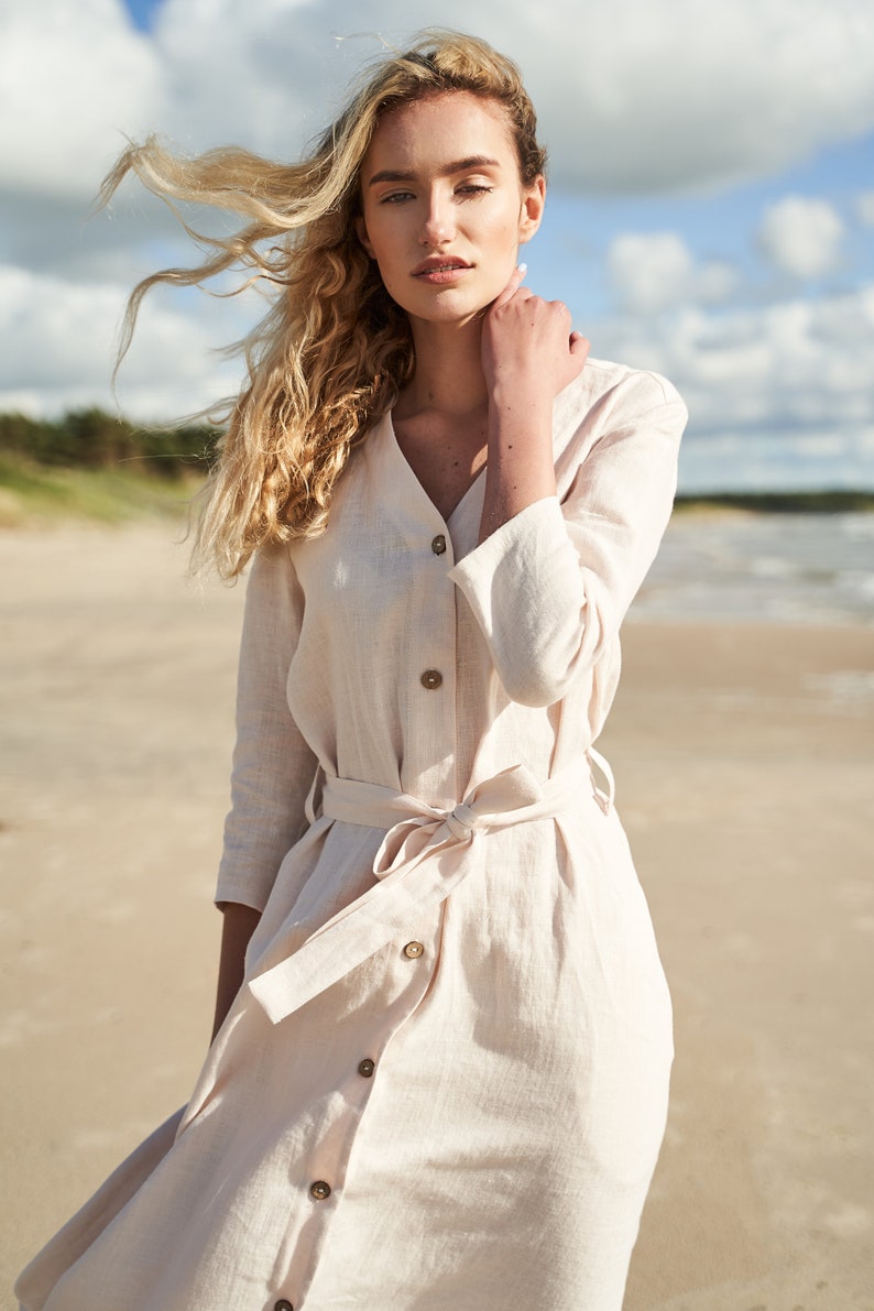 May include: A woman wearing a light beige linen dress with a button-up front and a tie belt. The dress has a v-neck and 3/4 sleeves. The woman is standing on a sandy beach with her hair blowing in the wind.