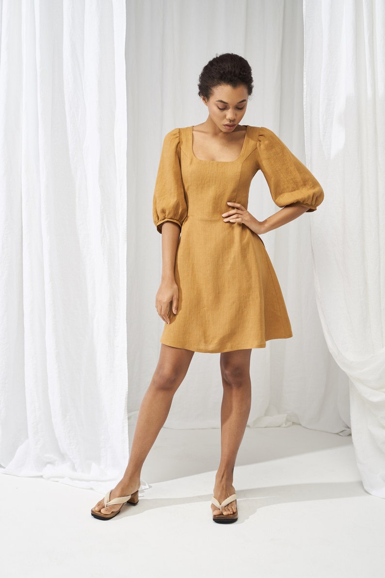 May include: A woman wearing a mustard yellow linen dress with puffed sleeves and a square neckline. She is standing in front of a white backdrop and wearing beige sandals.
