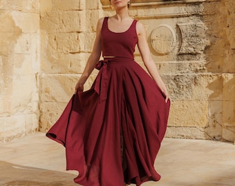May include: A woman wearing a burgundy linen top and skirt set. The skirt is a maxi skirt with a wrap style and a tie at the waist. The top is a sleeveless crop top with a scoop neck.