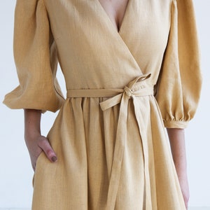 Puede incluir: Vestido cruzado amarillo mostaza con escote en V y cintura anudada. El vestido presenta mangas abullonadas hasta el codo y una falda acampanada. La modelo tiene una mano en el bolsillo del vestido. El vestido est&aacute; hecho de tela texturizada.