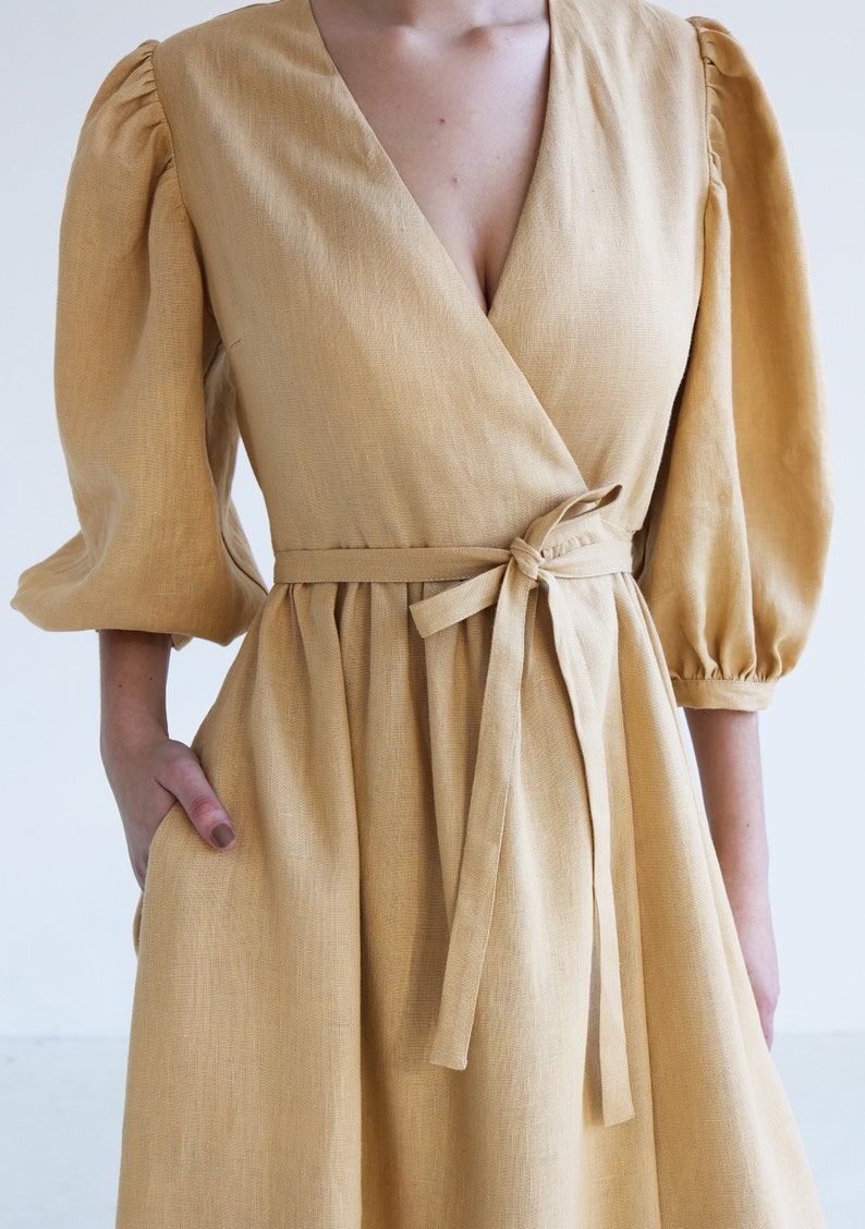 Puede incluir: Vestido cruzado amarillo mostaza con escote en V y cintura anudada. El vestido presenta mangas abullonadas hasta el codo y una falda acampanada. La modelo tiene una mano en el bolsillo del vestido. El vestido est&aacute; hecho de tela texturizada.