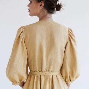 Puede incluir: Vestido de lino color beige claro con escote en V y mangas abullonadas voluminosas. El vestido tiene una cintura ce&ntilde;ida con un cintur&oacute;n a juego y una falda fluida. La tela tiene una textura sutil y el vestido parece estar hecho de un material ligero.