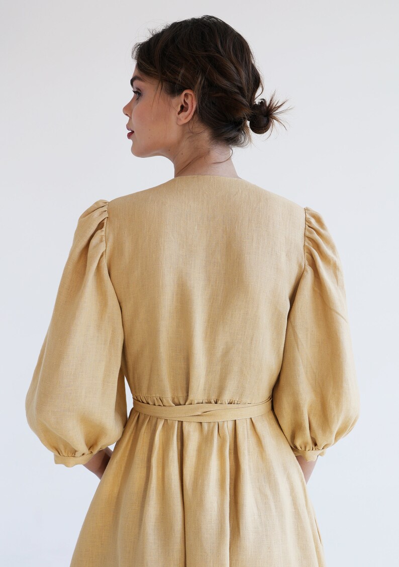 Puede incluir: Vestido de lino color beige claro con escote en V y mangas abullonadas voluminosas. El vestido tiene una cintura ce&ntilde;ida con un cintur&oacute;n a juego y una falda fluida. La tela tiene una textura sutil y el vestido parece estar hecho de un material ligero.