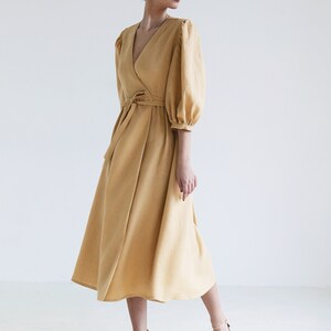 Puede incluir: Un vestido cruzado amarillo mostaza con escote en V y mangas abullonadas hasta el codo. El vestido presenta un cintur&oacute;n a juego y una falda fluida. La modelo lleva sandalias rosas.