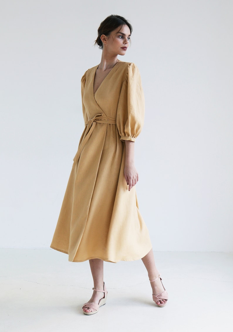 Puede incluir: Un vestido cruzado amarillo mostaza con escote en V y mangas abullonadas hasta el codo. El vestido presenta un cintur&oacute;n a juego y una falda fluida. La modelo lleva sandalias rosas.