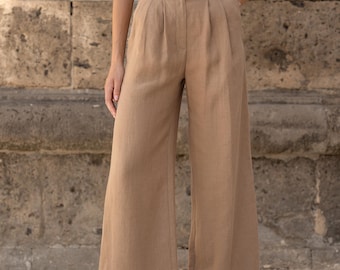 Pode incluir: Calça de linho bege de perna larga com cinto de couro castanho e fivela prateada. A calça tem pregas na cintura e um caimento solto.