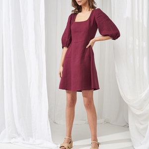 May include: A burgundy linen dress with a square neckline and puffed sleeves. The dress has a fitted bodice and a flowy skirt. The model is wearing the dress with beige platform sandals.