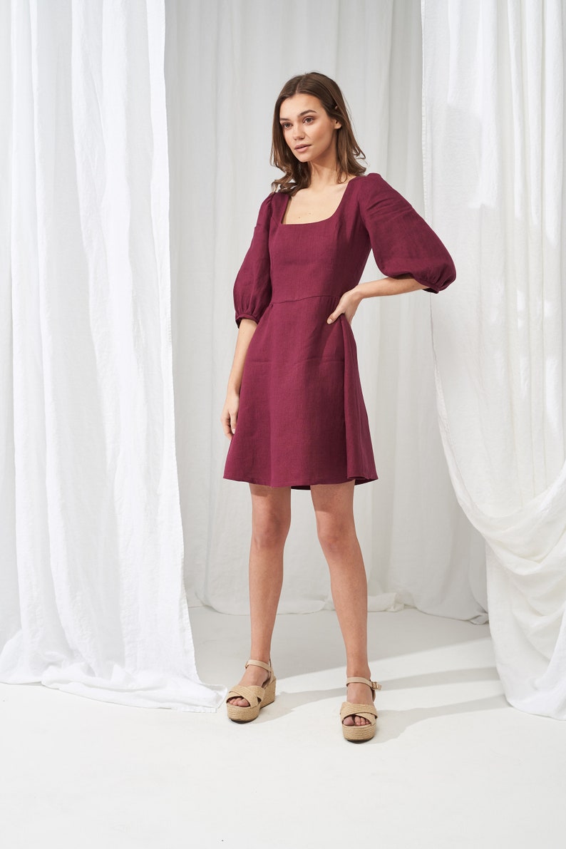 May include: A burgundy linen dress with a square neckline and puffed sleeves. The dress has a fitted bodice and a flowy skirt. The model is wearing the dress with beige platform sandals.