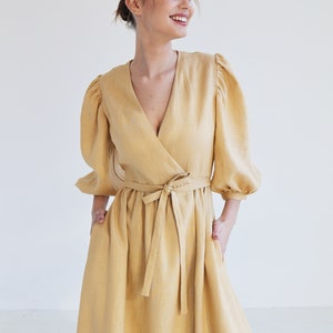 Puede incluir: Un vestido cruzado amarillo mostaza con escote en V y cintur&oacute;n anudado. El vestido presenta mangas abullonadas de tres cuartos y una falda fluida. La persona lleva el vestido y tiene las manos en los bolsillos.
