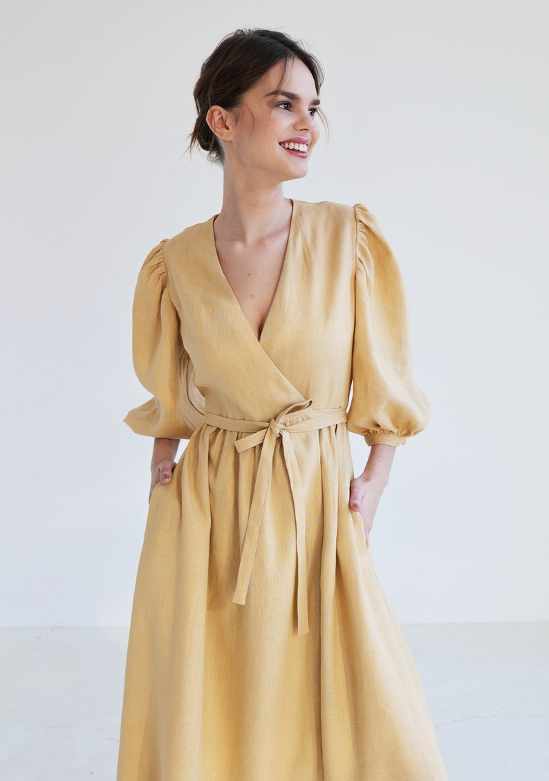 Puede incluir: Un vestido cruzado amarillo mostaza con escote en V y cintur&oacute;n anudado. El vestido presenta mangas abullonadas de tres cuartos y una falda fluida. La persona lleva el vestido y tiene las manos en los bolsillos.