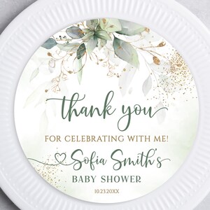 May include: A white plate with a decorative insert featuring a floral design and the words "thank you" in green script. The insert includes the text "FOR CELEBRATING WITH ME! Sofia Smith's BABY SHOWER 10.23.20XX". Gold cutlery is placed to the right.