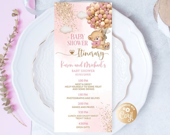 May include: A pink baby shower itinerary card with gold text and a teddy bear illustration holding balloons. The card includes the text "Baby Shower Itinerary" and the names "Karen and Michael's". The card is on a white plate.