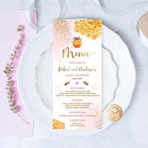 May include: A baby shower menu card with a watercolor design. The menu features a honeybee theme with a jar of honey and honeycomb illustrations. The menu includes appetizers, entrees, a momosa bar, and a dessert bar. The menu is on a white plate.