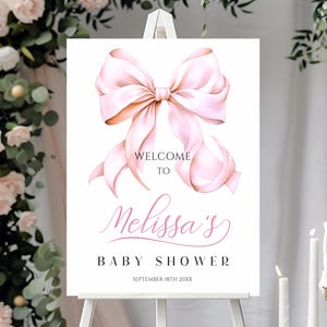 May include: A white rectangular welcome sign featuring a large, soft pink watercolor bow at the top. The text reads: WELCOME TO, Melissa's, BABY SHOWER, SEPTEMBER 18TH 20XX. The sign is displayed on a white easel stand against a floral background.