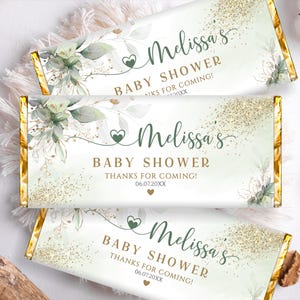 Op de afbeelding: Drie baby shower candybar wrappers met een goud en wit ontwerp. De wrappers bevatten de tekst "Melissa's BABY SHOWER" in groen schrift, met bloemaccenten en gouden glitter.