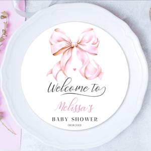 Puede incluir: Una pegatina redonda blanca con un lazo rosa y el texto "Bienvenido al Baby Shower de Melissa 08.08.20XX".