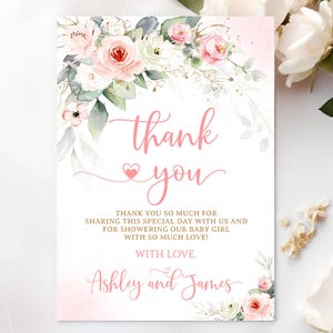 May include: A thank you card with a floral design. The card features pink roses, white flowers, and green leaves. The words "thank you" are written in pink script, with additional text below. The card is for a baby girl.