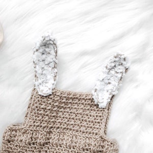 May include: A close-up of a crochet bunny romper in a neutral beige color. The romper features two fluffy white and gray bunny ears attached to the top. The crochet stitch creates a textured surface.