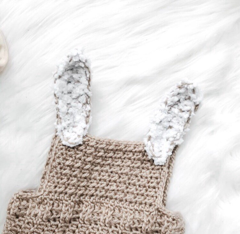 May include: A close-up of a crochet bunny romper in a neutral beige color. The romper features two fluffy white and gray bunny ears attached to the top. The crochet stitch creates a textured surface.