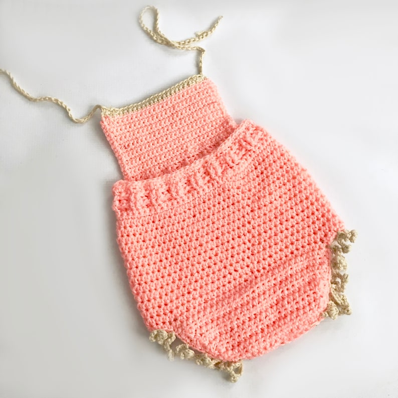 May include: A handmade, peach-colored crochet romper with a cream-colored trim and straps. The romper features a textured stitch pattern and a scalloped edge. The garment is designed for a baby or toddler.