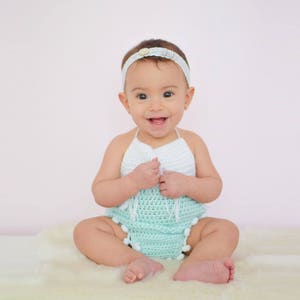 May include: A baby girl wearing a white and light green crocheted outfit with a white headband. The baby is sitting on a white fluffy rug and smiling at the camera.