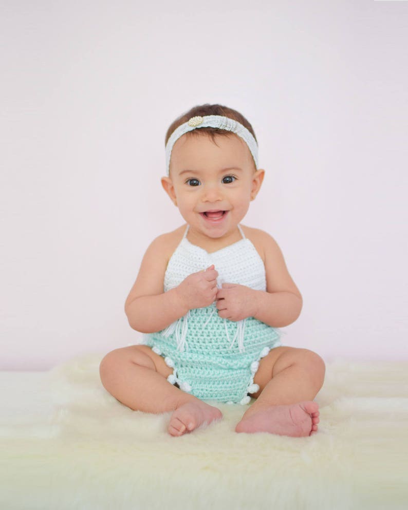 May include: A baby girl wearing a white and light green crocheted outfit with a white headband. The baby is sitting on a white fluffy rug and smiling at the camera.