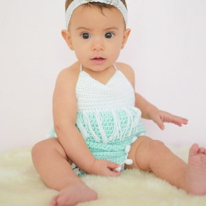 May include: A baby girl wearing a light green and white crocheted halter top with fringe and white pom-poms. She is wearing a white headband with a pearl accent.