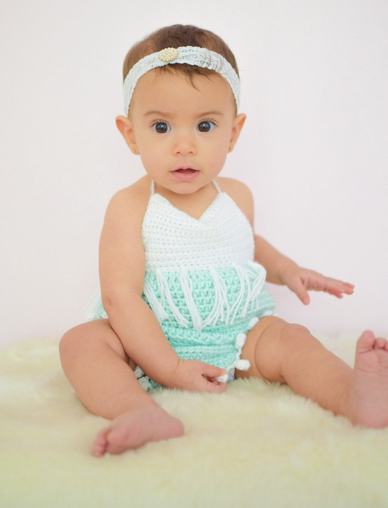 May include: A baby girl wearing a light green and white crocheted halter top with fringe and white pom-poms. She is wearing a white headband with a pearl accent.