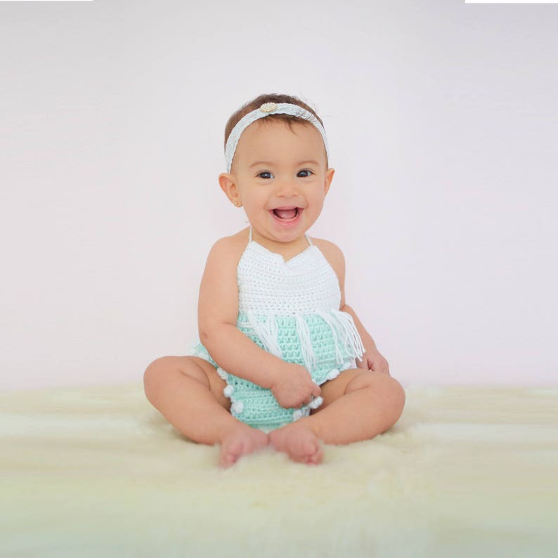 May include: A baby girl wearing a light green crocheted halter top with white trim and white pom-poms. She is sitting on a white fluffy surface and smiling.