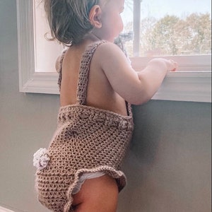 May include: A light brown crochet baby romper with a bunny tail detail. The romper has shoulder straps and a ruffled edge. The baby is looking out a window.