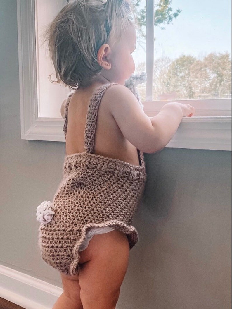 May include: A light brown crochet baby romper with a bunny tail detail. The romper has shoulder straps and a ruffled edge. The baby is looking out a window.