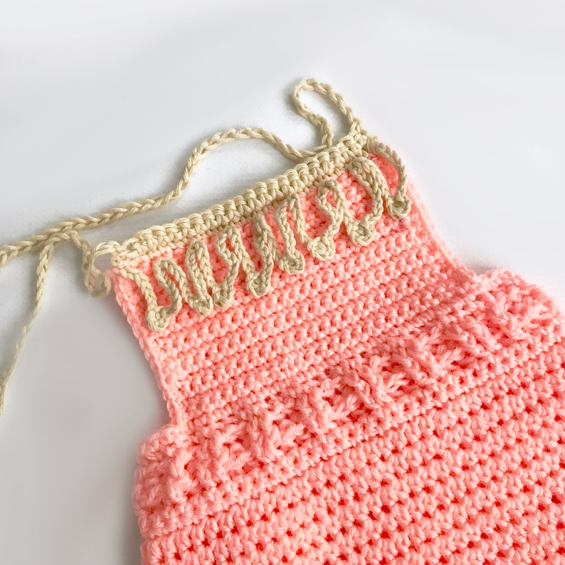 May include: A handmade, light pink crochet top with a decorative beige trim along the neckline. The top features a textured stitch pattern and delicate straps. This item is perfect for a baby or toddler.