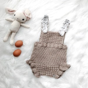 May include: A tan crochet baby romper with bunny ears, two brown eggs, and a stuffed bunny toy on a white fluffy surface. The romper has a textured stitch and ruffled edges. The bunny toy is cream-colored with pink accents.