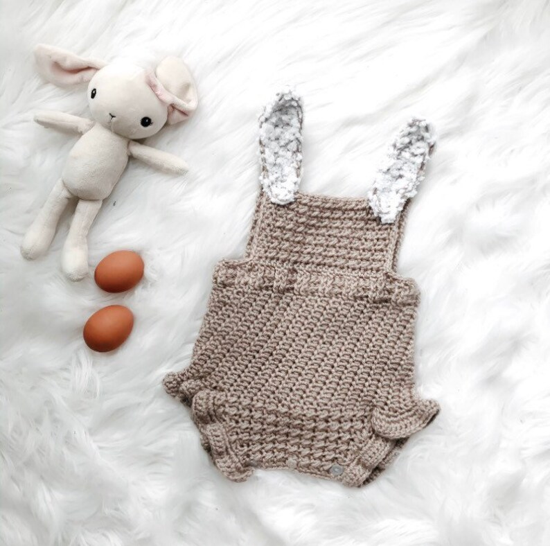 May include: A tan crochet baby romper with bunny ears, two brown eggs, and a stuffed bunny toy on a white fluffy surface. The romper has a textured stitch and ruffled edges. The bunny toy is cream-colored with pink accents.