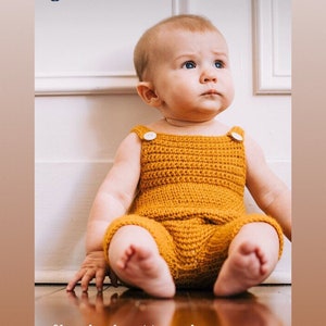 May include: A baby wearing a crocheted yellow jumpsuit with white buttons. The baby is sitting on a wooden floor and looking up.