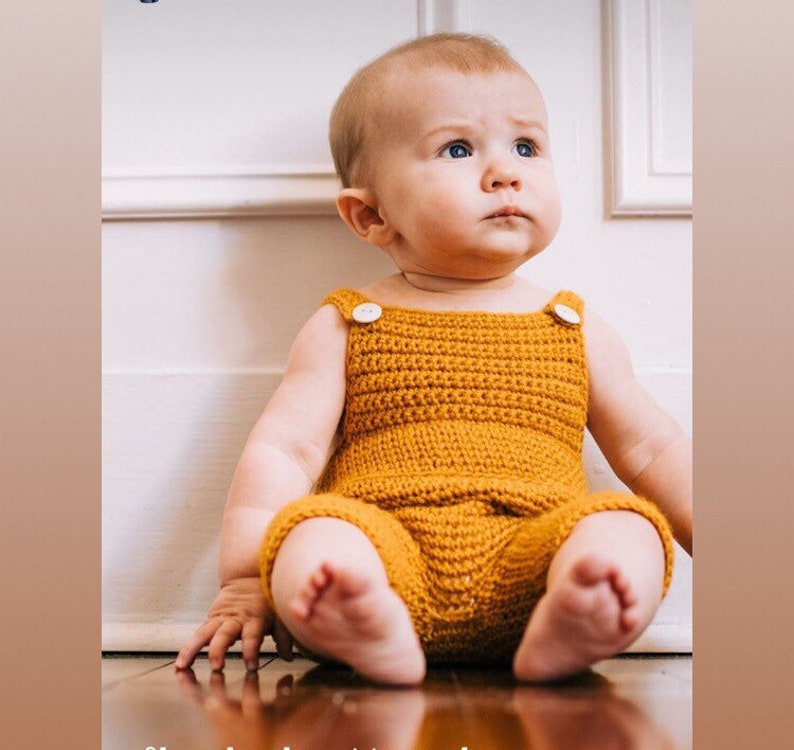 May include: A baby wearing a crocheted yellow jumpsuit with white buttons. The baby is sitting on a wooden floor and looking up.