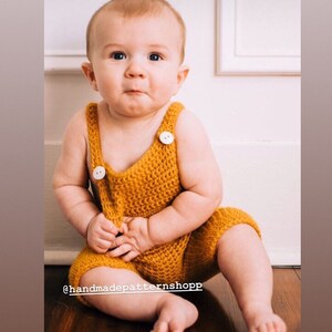 May include: A baby wearing a crocheted yellow romper with white buttons. The romper has a sleeveless design and a wide leg. The baby is sitting on a wooden floor.